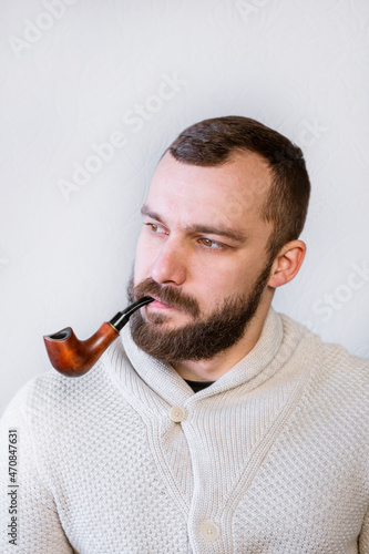 Smiling man gentleman with beard and mustache in light cardigan holds and smokes pipe, stylish fancy trendy man