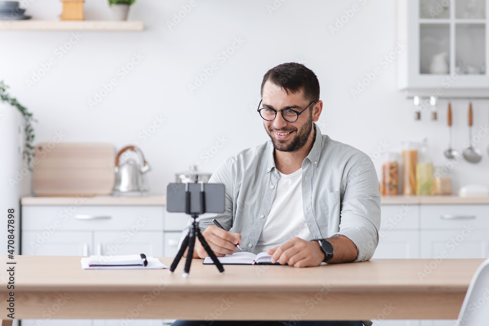 Cheerful mature caucasian male in glasses makes notes and looks at smartphone webcam make video call