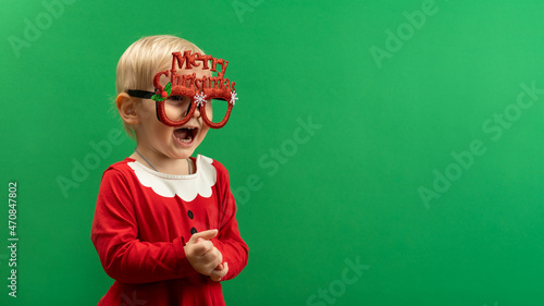 Toddler girl in red Christmas costume and glasses laughs on green background COPYSPACE