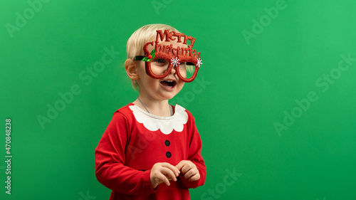  Toddler girl in red Christmas costume and glasses laughs on green background COPYSPACE