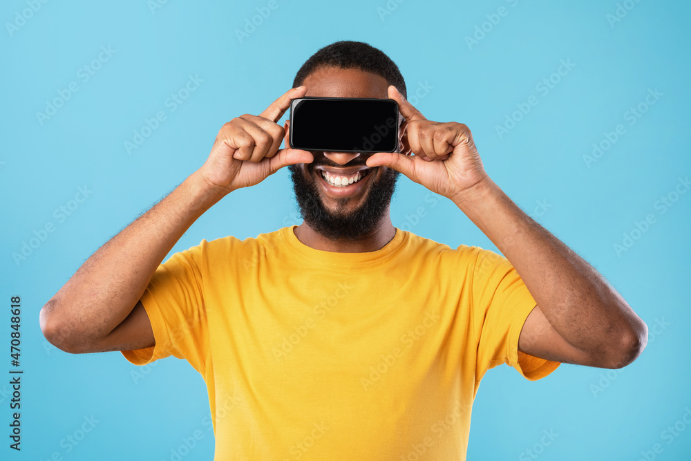 Fototapeta premium Happy black guy covering eyes with blank smartphone, ofering mockup for website or application on blue studio background