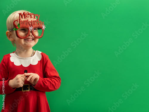  Happy Toddler girl in red Christmas costume and glasses laughs on green background COPYSPACE