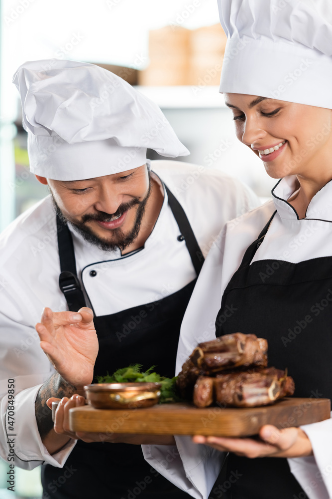 Smiling asian chef showing okay gesture near colleague with roasted meat Smiling asian chef showing okay gesture near colleague with roasted meat