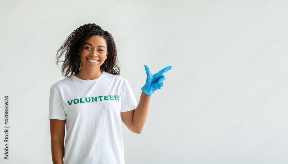 Look here. Young african american volunteer in gloves pointing aside at empty space, standing over light studio wall Look here. Young african american volunteer in gloves pointing aside at empty space, standing over light studio wall