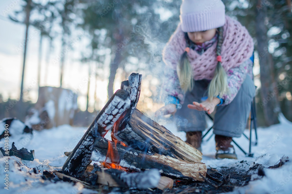 Obraz premium The girl in winter clothes warms her hands by the fire in snow-covered forest, winter trip at weekend. Family winter picnic, active lifestyle, focus on woods
