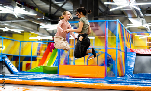 Pretty girls jumping together on colorful trampoline at playground park. Two sisters having fun during active entertaiments indoor photo