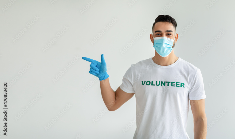 Look there. Male volunteer in medical mask pointing aside showing free space while standing over light studio wall Look there. Male volunteer in medical mask pointing aside showing free space while standing over light studio wall