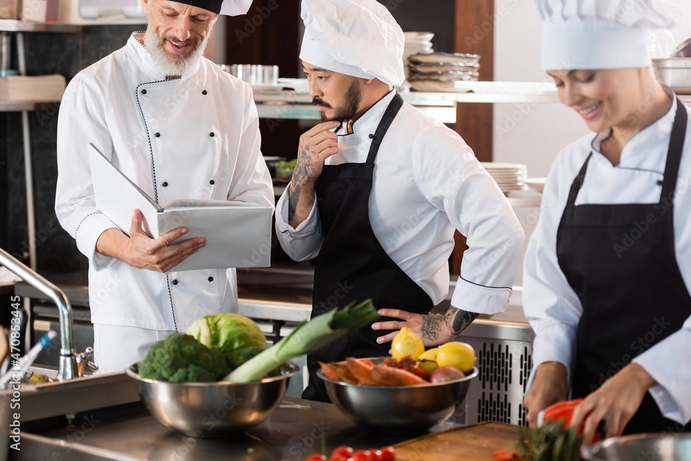 Smiling chef holding cookbook near asian colleague and vegetables in kitchen Smiling chef holding cookbook near asian colleague and vegetables in kitchen