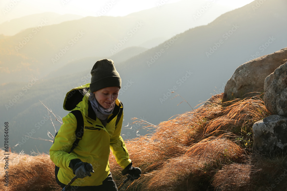 young woman with hiking sticks, wearing bright green windproof jacket, big backpack, hat, going up in the rocky mountains covered with stones and long dry grass