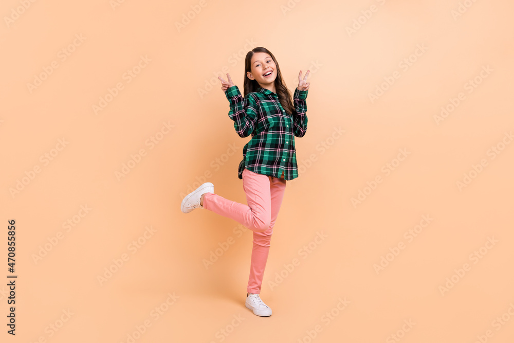 Photo of excited funny school girl dressed checkered clothes smiling walking showing tow v-signs isolated beige color background Photo of excited funny school girl dressed checkered clothes smiling walking showing tow v-signs isolated beige color background