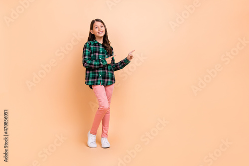 Photo of adorable sweet school girl dressed checkered clothes smiling pointing two fingers empty space isolated beige color background