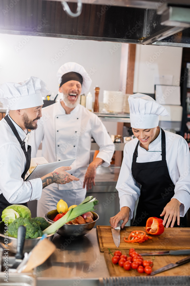 Asian chef holding cookbook near laughing colleagues and fresh food in kitchen Asian chef holding cookbook near laughing colleagues and fresh food in kitchen