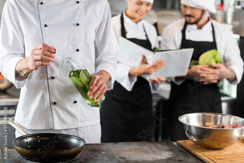 Chef holding bottle of olive oil near frying pan and blurred colleagues with cookbook in kitchen