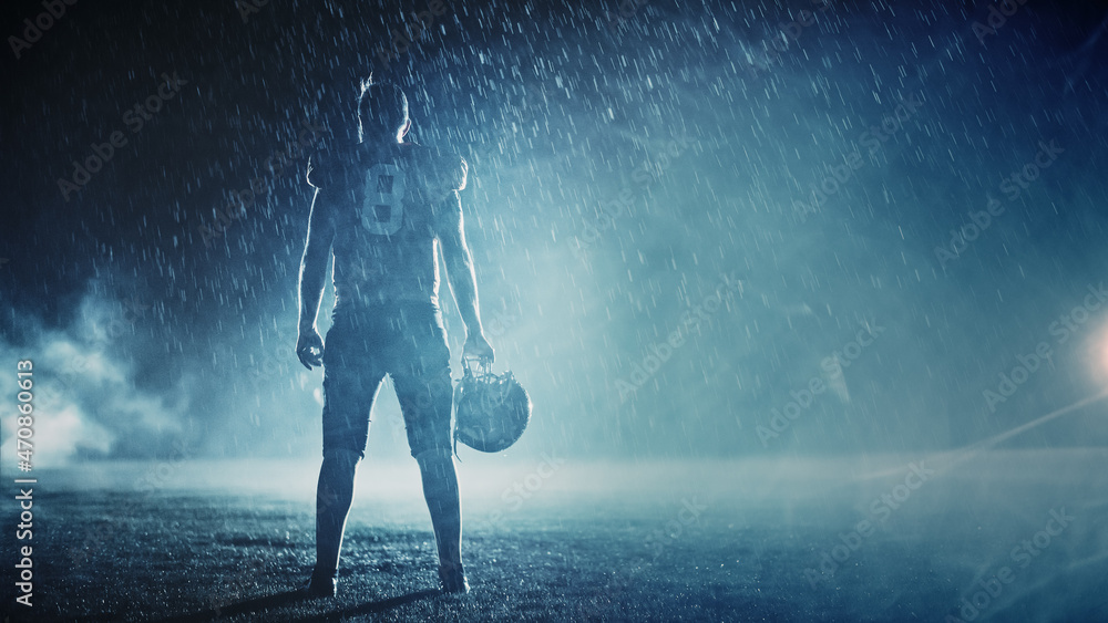 Fototapeta premium American Football Field: Lonely Athlete Warrior Standing on a Field Holds his Helmet and Ready to Play. Player Preparing to Run, Attack and Score Touchdown. Rainy Night with Dramatic Fog, Blue Light
