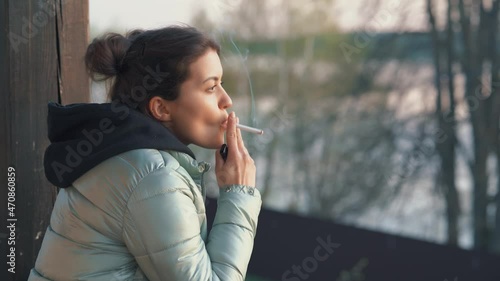 A beautiful young woman smokes a cigarette in the evening on the veranda outdoors in cool weather in the evening. Close-up of a smoking girl.