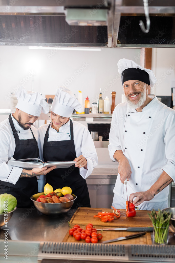 Smiling mature chef cutting bell pepper near interracial colleagues reading cookbook in kitchen Smiling mature chef cutting bell pepper near interracial colleagues reading cookbook in kitchen