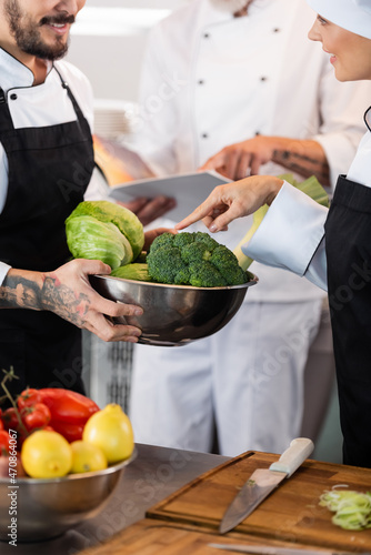 Smiling chef pointing at vegetables in bowl near colleague in kitchen