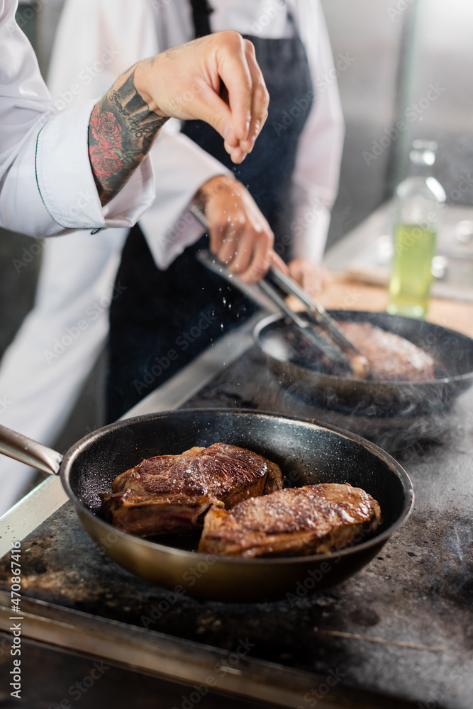 Cropped view of tattooed chef pouring salt while roasting meat on cooktop in kitchen Cropped view of tattooed chef pouring salt while roasting meat on cooktop in kitchen