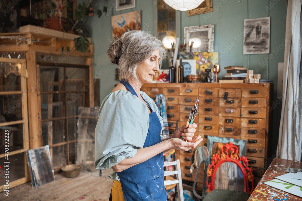 Female artist in apron holding brushes and standing at her studio before drawing