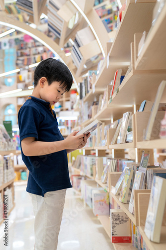 Young Asia boy standing in the modern and nice interior bookstore. Choosing and holding a book. Many bookshelf in the shop. Kid learning and education concept. Colorful book at library