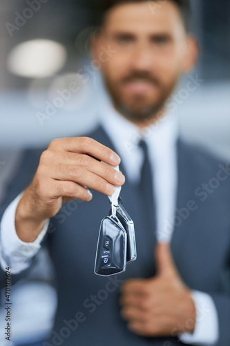 Blur background of caucasian male customer standing at car salon, holding keys from new car and showing thumb up. Focus on male hands with keys. 