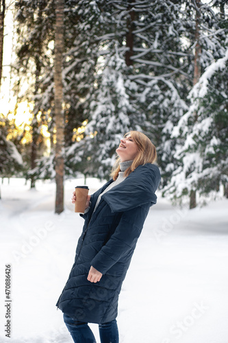 A nice middle-aged blonde woman walks through the winter forest with a cup of hot drink. Winter activity and healthy lifestyle.