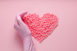 Pink heart-shaped wax granules for depilation on a pink background and the hand of a master in a pink glove. Epilation, depilation, removal of unwanted hair with wax. Top view.