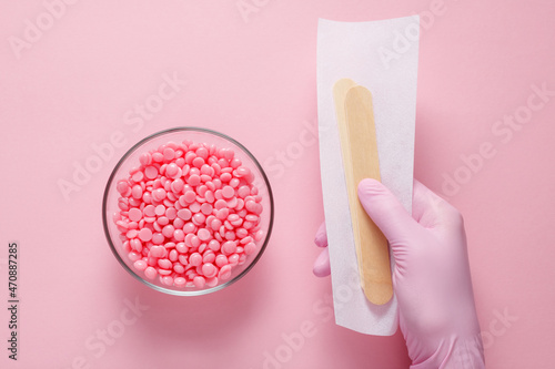 Pink granules of wax for depilation in a glass bowl on a pink background and a hand of a master in a pink glove holding a strip for depilation and wooden spatulas. Epilation, depilation photo