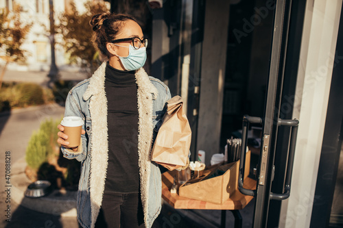 Young woman wearing protective face mask holding takeaway food and coffee to go, standing near restaurant window. Social distancing during quarantine caused pandemic.