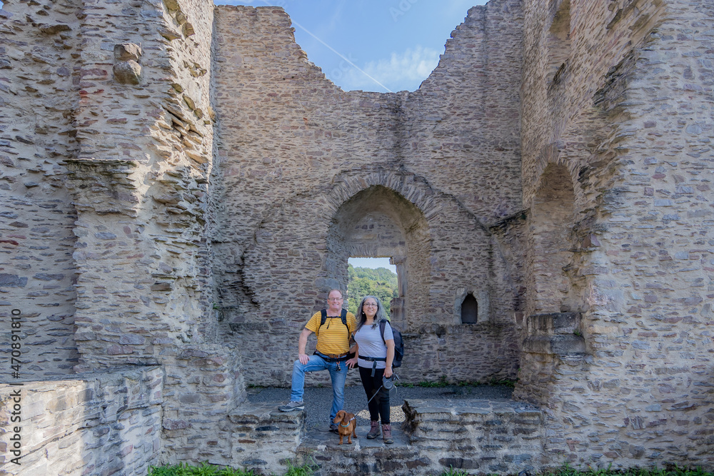 Mature tourist couple smiling and looking at the camera next to their brown dog, Brandenburg castle in ruin with stone walls, window in the background, sunny summer day in Luxembourg