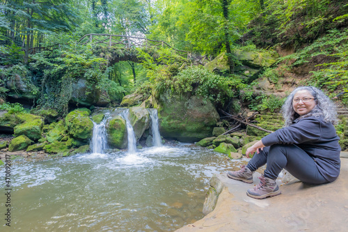 Female hiker tourist sitting on rock, Schiessentümpel waterfall on Black Ernz river and abundant green wild vegetation in background, greyish hair, looking at camera, Mullerthal Trail, Luxembourg photo