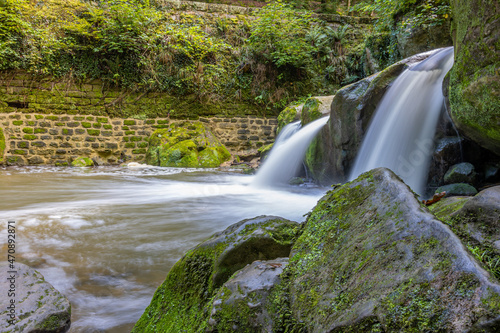 Side view of the Schiessentümpel waterfall with water flowing between the rocks in the Black Ernz river, brick wall with moss in the background, Mullerthal Trail, Luxembourg. Long exposure image photo