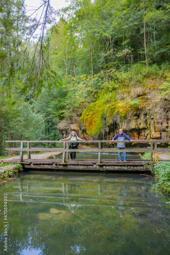 Mullerthal Trail, tourist couple standing on a wooden bridge over the basin, Kallektuffquell waterfall, water flowing between rock formations with green moss in the background, sunny day in Luxembourg