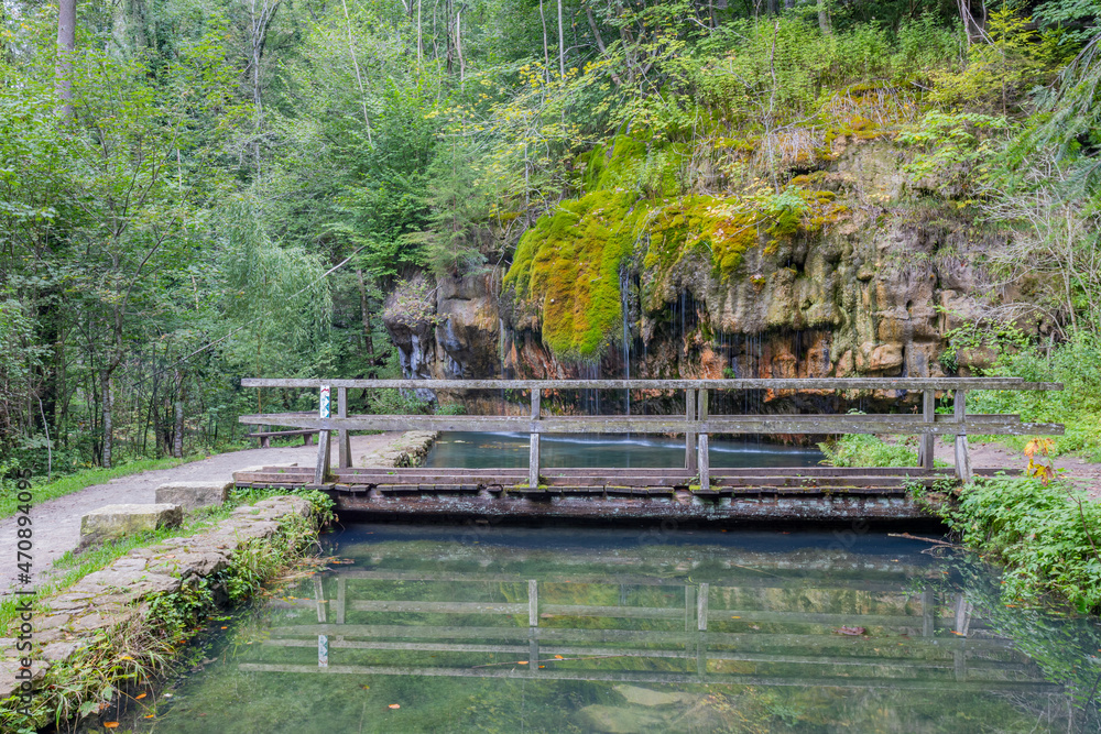 Wooden bridge over the basin with reflection on the water surface, Kallektuffquell waterfall with water flowing between rock formations with green moss in the background, sunny day in Luxembourg Wooden bridge over the basin with reflection on the water surface, Kallektuffquell waterfall with water flowing between rock formations with green moss in the background, sunny day in Luxembourg