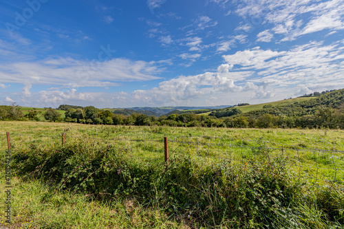 Landscape of the countryside with its hills covered with wild green grass, lush trees, sunny day with a stunning blue sky and white clouds in the background, Luxembourg