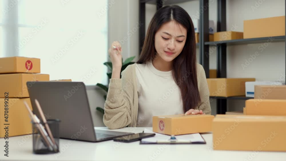 Young asian business woman writing address to sending product to customer using smartphone scan qr code on the box. Online business working at home. Startup Small business owner entrepreneur Young asian business woman writing address to sending product to customer using smartphone scan qr code on the box. Online business working at home. Startup Small business owner entrepreneur