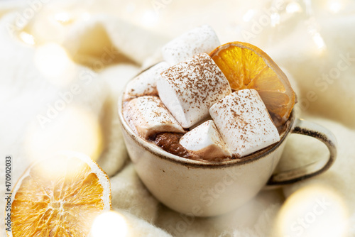 Traditional Christmas cocoa in a ceramic cup with marshmallows and orange chips on a light background with bokeh