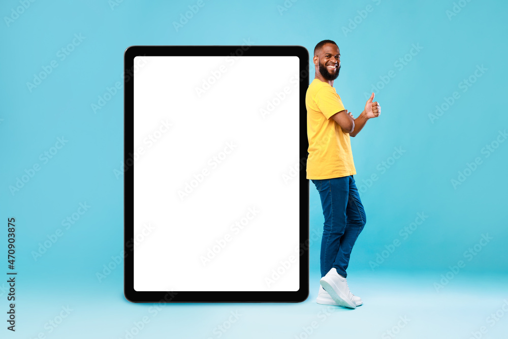 Positive black guy showing thumb up gesture next to huge digital tablet on blue studio background, mockup Positive black guy showing thumb up gesture next to huge digital tablet on blue studio background, mockup