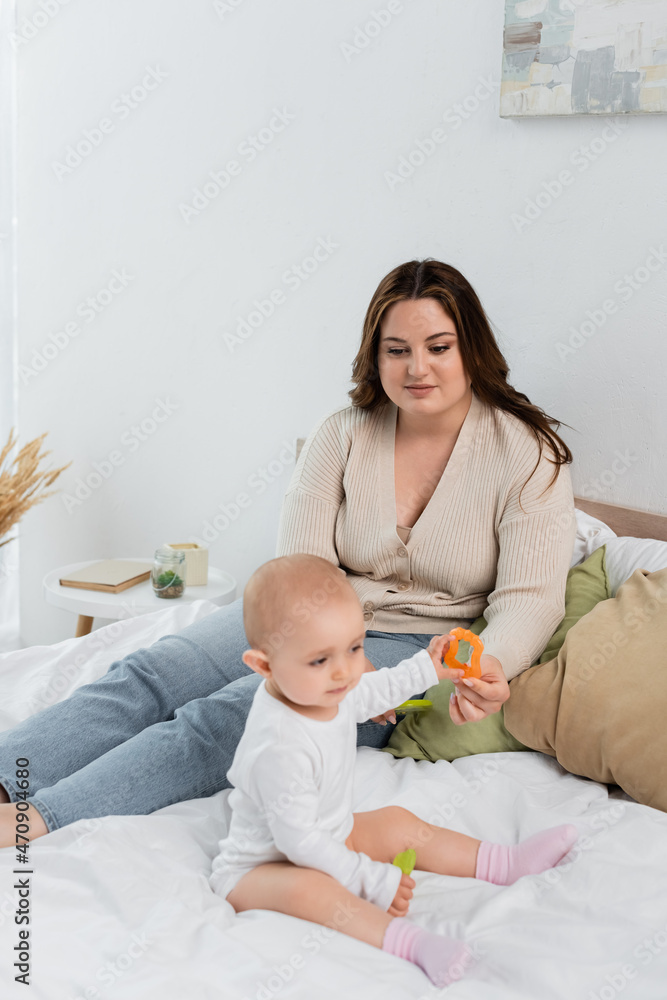 Young plus size woman looking at baby daughter with toys on bed.