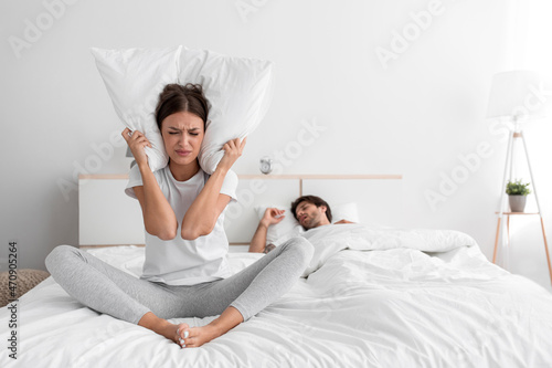 Stressed sad millennial caucasian lady sits on bed covers ears with pillow and suffers from snoring of sleeping man photo