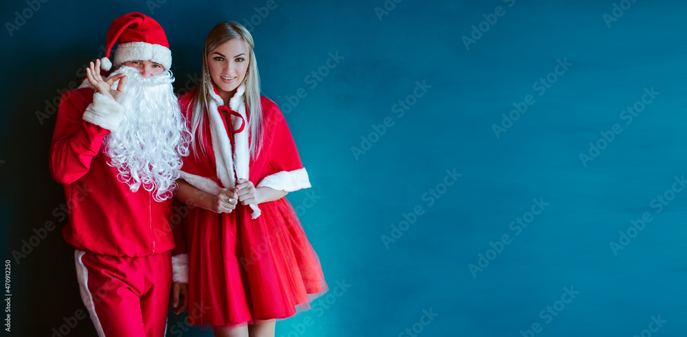 funny and mischievous, trendy santa claus with a beard and a red winter dress, with his assistant shows sophisticated signs, inviting to the party time on a blue background with a place for display an