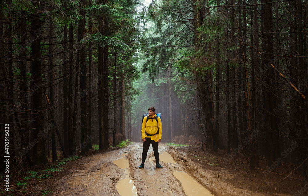 Male hiker stands on a winding road in the mountains with a backpack on his back against the backdrop of a beautiful misty forest after the rain. Male hiker stands on a winding road in the mountains with a backpack on his back against the backdrop of a beautiful misty forest after the rain.
