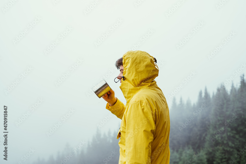 Male hiker in a yellow jacket in the mountains drinks tea from a metal cup on a background of misty mountain forest.