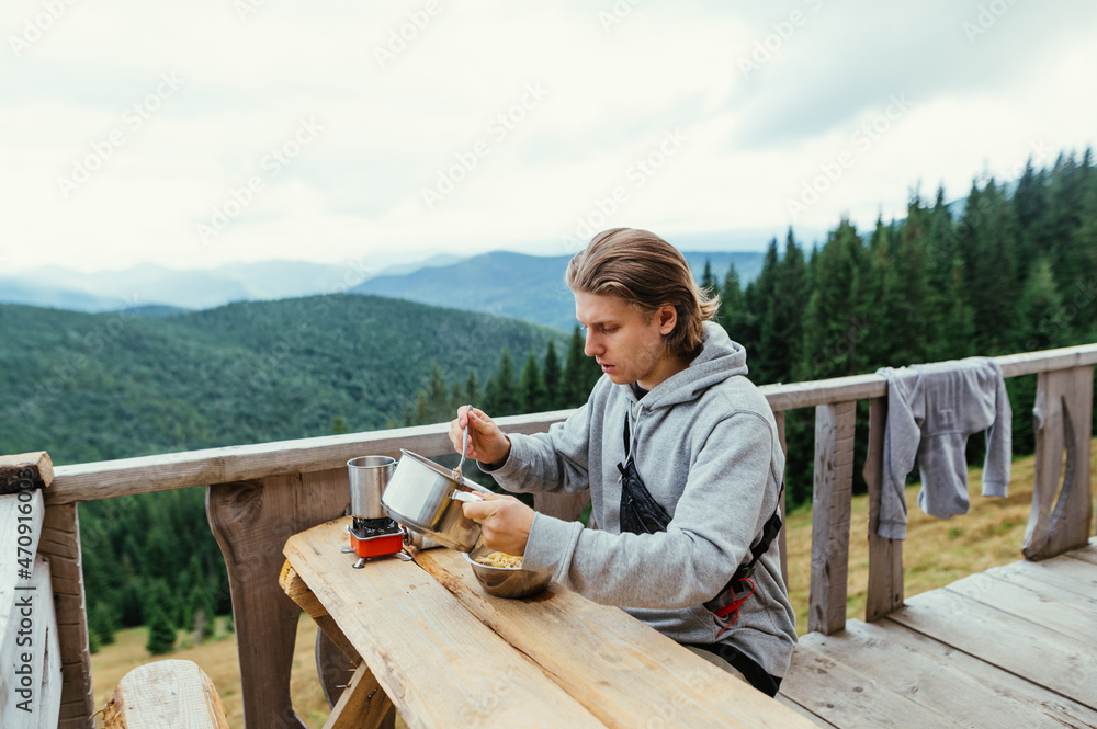 Fototapeta premium Guy tourist lunch on the terrace of a wooden bungalow on a background of mountains. The tourist spent the night in a mountain house, eating pasta for breakfast.