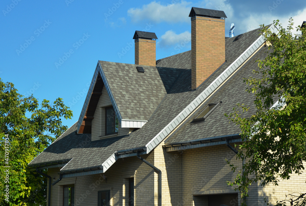 A house with a pitched roofing construction with asphalt shingles installed, attic windows, skylights, soffit and fascia boards, ventilation, two brick chimneys, and roof gutters. A house with a pitched roofing construction with asphalt shingles installed, attic windows, skylights, soffit and fascia boards, ventilation, two brick chimneys, and roof gutters.