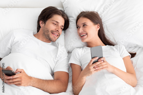Happy caucasian millennial wife and husband looking at smartphone, lying on bed in white bedroom interior, top view