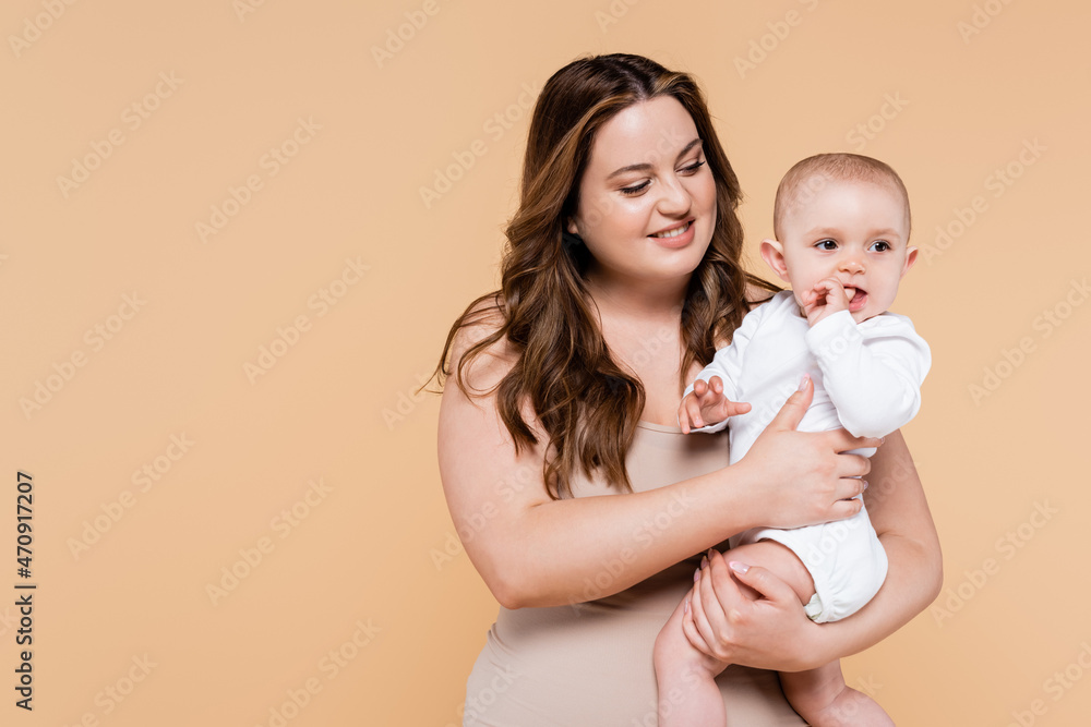 Smiling body positive mother holding baby with finger near mouth isolated on beige. Smiling body positive mother holding baby with finger near mouth isolated on beige.