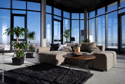 Fashionable modern apartment with panoramic windows  grey comfy couch  coffee table and green plants in pots. Stylish interior of flat.
