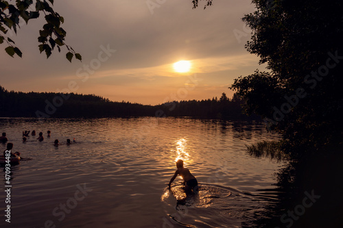 Silhouettes of people swimming in evening lake in reflection of sunset among the trees