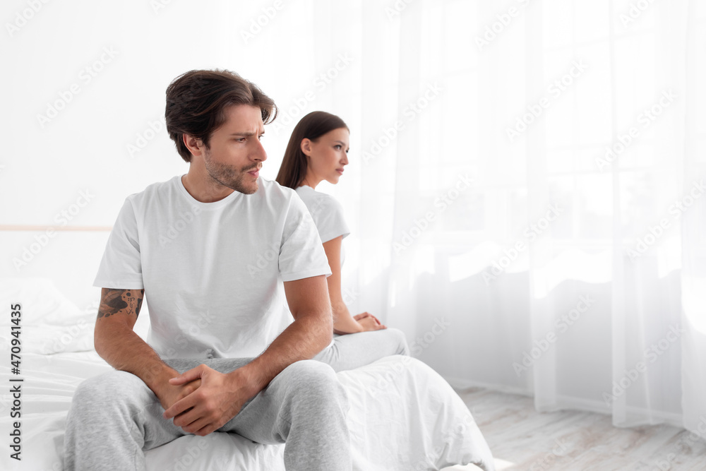 Unhappy upset offended caucasian millennial husband ignores wife sitting in bed in white bedroom interior Unhappy upset offended caucasian millennial husband ignores wife sitting in bed in white bedroom interior
